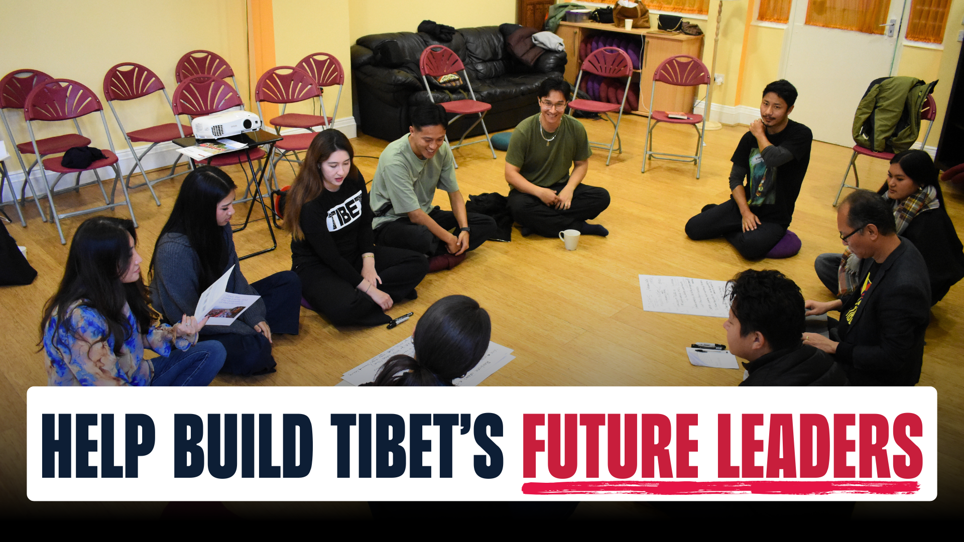 A group of a dozen young Tibetans sit cross legged on the floor in a circle in a room with wooden floors. In the young people’s hands are information sheets regarding the campaigning workshop. In the centre of the circle are several poster papers with ide